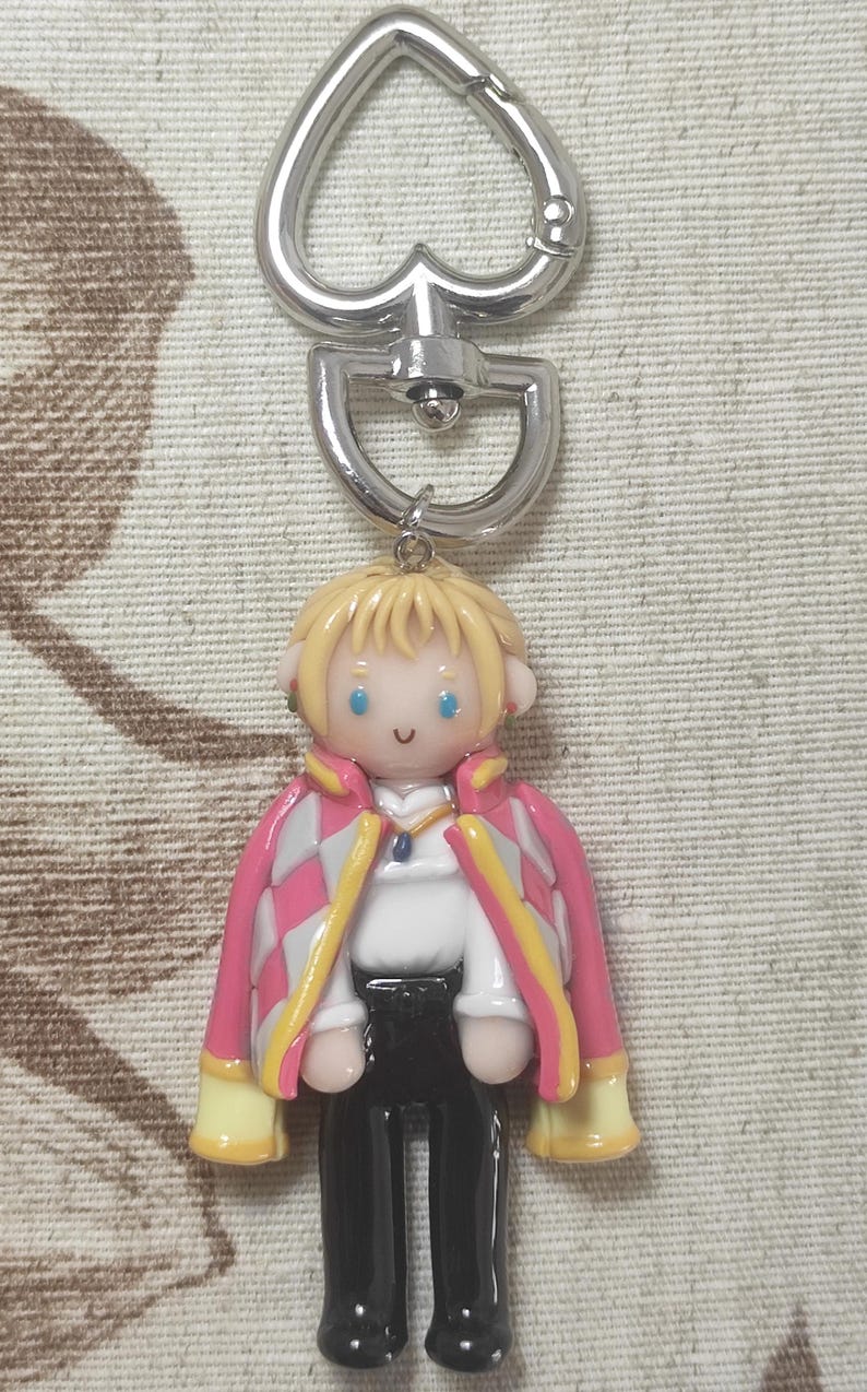 May include: A keychain featuring a cartoon figure with blonde hair, blue eyes, and a pink and white checkered jacket with yellow accents. The figure is wearing a white shirt and black pants. The keychain has a silver heart-shaped clasp.
