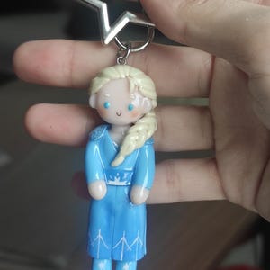 May include: A blue polymer clay keychain featuring a character with blonde hair in a side braid, wearing a blue dress with white accents. The keychain has a silver star-shaped top and a silver ring.