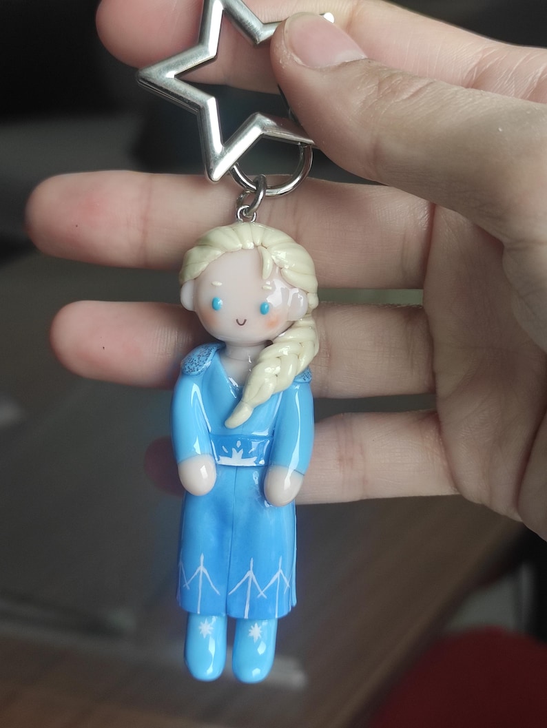 May include: A blue polymer clay keychain featuring a character with blonde hair in a side braid, wearing a blue dress with white accents. The keychain has a silver star-shaped top and a silver ring.