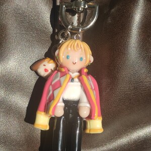 May include: A keychain featuring a miniature figurine of a character with blonde hair, blue eyes, and a pink and white jacket. The figure is attached to a silver-colored metal clasp. A small brown and white figure is attached to the side of the main figure.