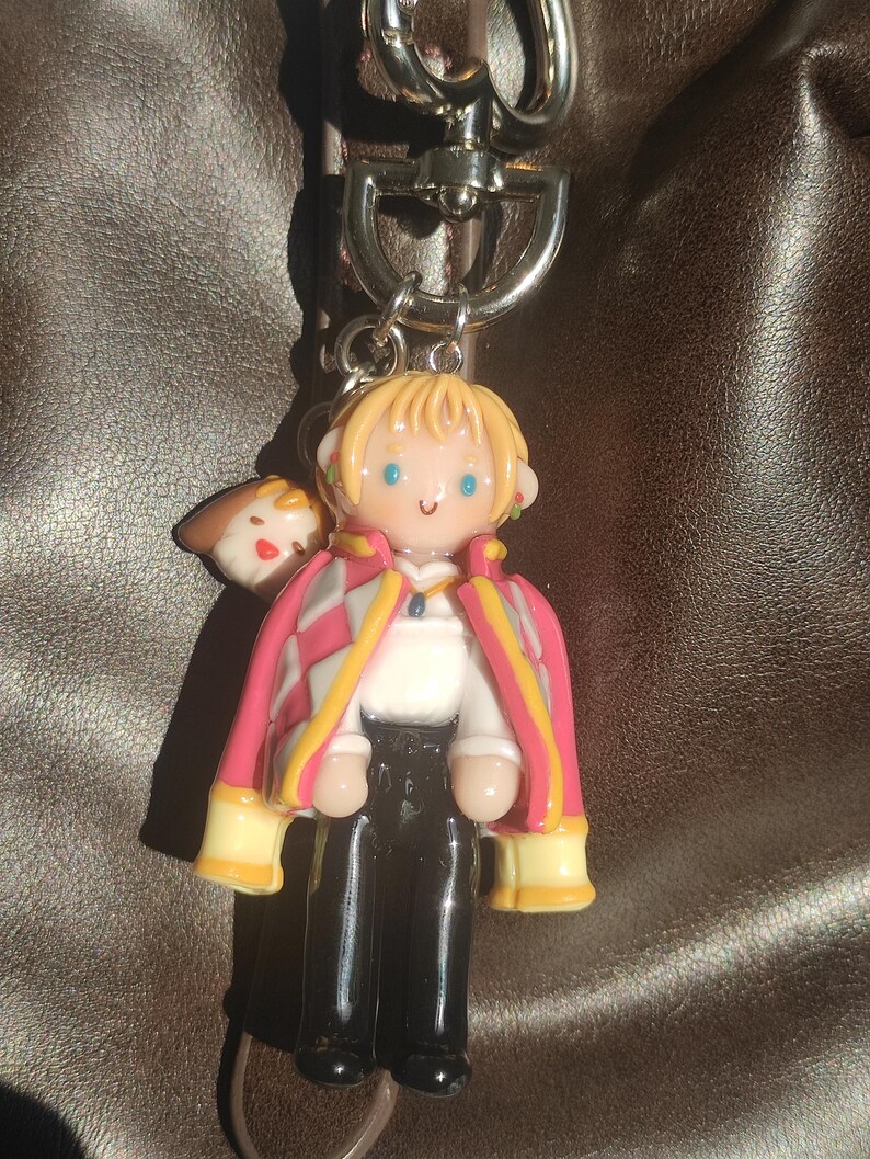 May include: A keychain featuring a miniature figurine of a character with blonde hair, blue eyes, and a pink and white jacket. The figure is attached to a silver-colored metal clasp. A small brown and white figure is attached to the side of the main figure.