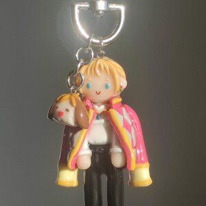 May include: A handmade keychain featuring a miniature figure of a character with blonde hair, wearing a pink and white jacket, black pants, and a white shirt. A small dog-like charm is attached. The keychain has a silver clasp.