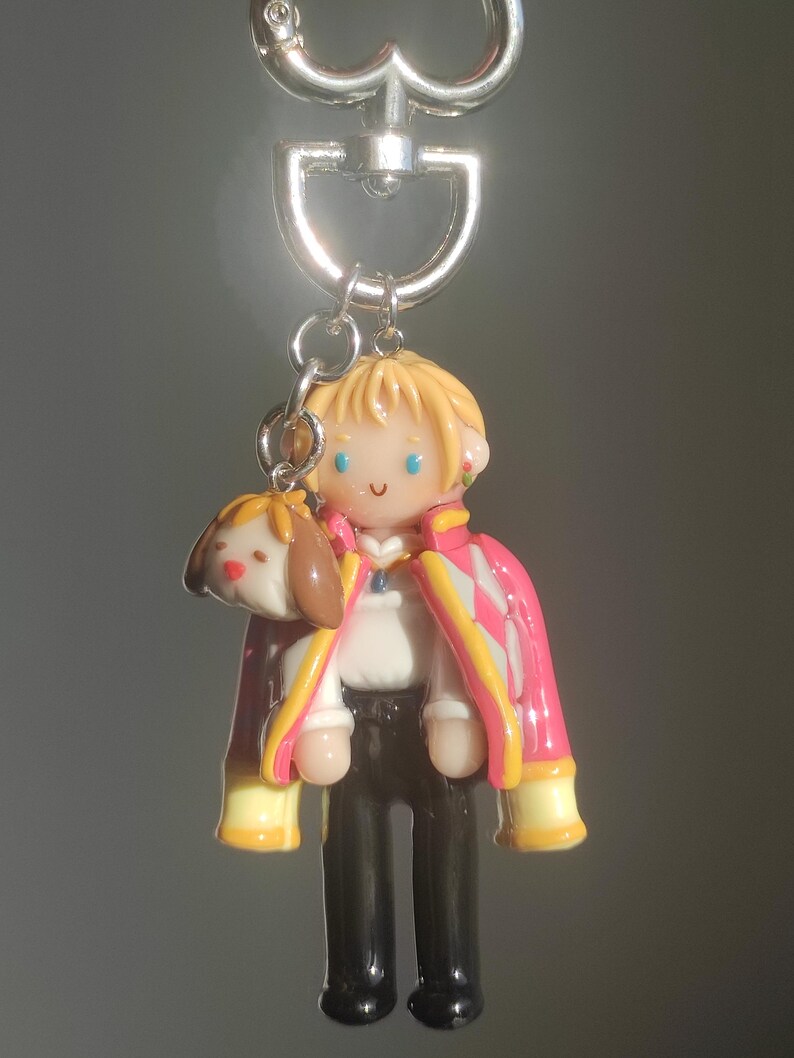 May include: A handmade keychain featuring a miniature figure of a character with blonde hair, wearing a pink and white jacket, black pants, and a white shirt. A small dog-like charm is attached. The keychain has a silver clasp.