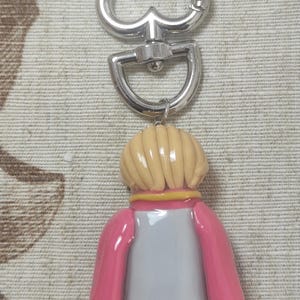 May include: A keychain featuring a figure with a light blonde hairstyle, wearing a pink jacket over a gray shirt and black pants. The keychain has a silver-colored heart-shaped clasp and a swivel connector.