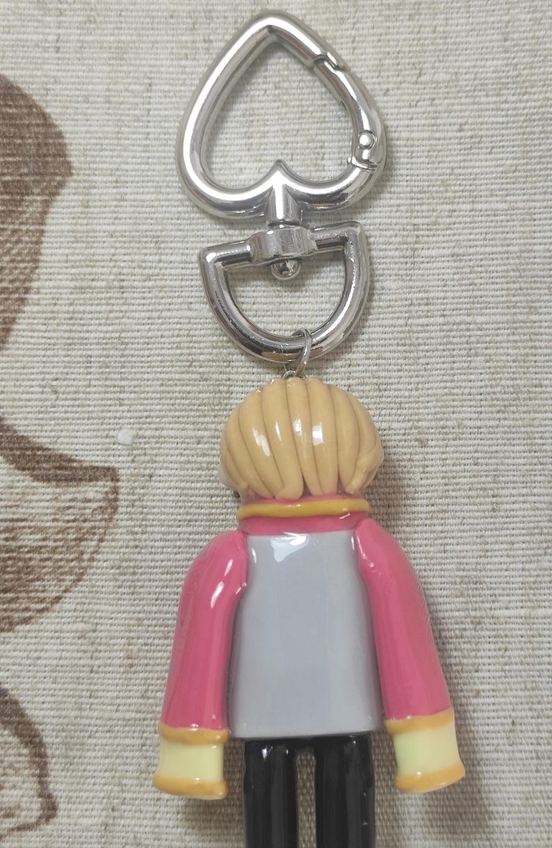 May include: A keychain featuring a figure with a light blonde hairstyle, wearing a pink jacket over a gray shirt and black pants. The keychain has a silver-colored heart-shaped clasp and a swivel connector.