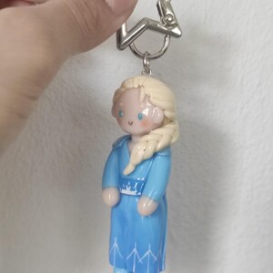 May include: A keychain featuring a miniature figurine of a character from a popular animated film. The figurine is dressed in a blue dress with white accents and has blonde hair. The keychain has a silver star-shaped clasp.