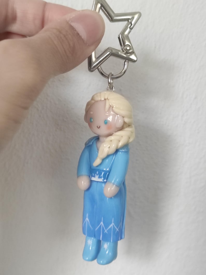 May include: A keychain featuring a miniature figurine of a character from a popular animated film. The figurine is dressed in a blue dress with white accents and has blonde hair. The keychain has a silver star-shaped clasp.