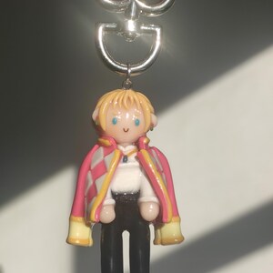 May include: A handmade keychain featuring a miniature figurine of a character with blonde hair, blue eyes, and a pink and white jacket. The keychain has a silver-colored metal clasp.