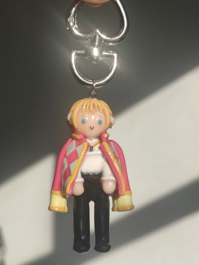 May include: A handmade keychain featuring a miniature figurine of a character with blonde hair, blue eyes, and a pink and white jacket. The keychain has a silver-colored metal clasp.