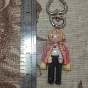 May include: A handmade keychain featuring a miniature figure of a character with blonde hair, blue eyes, and a pink and yellow jacket. The figure is attached to a silver-colored heart-shaped clasp. The figure is approximately 2 inches tall.