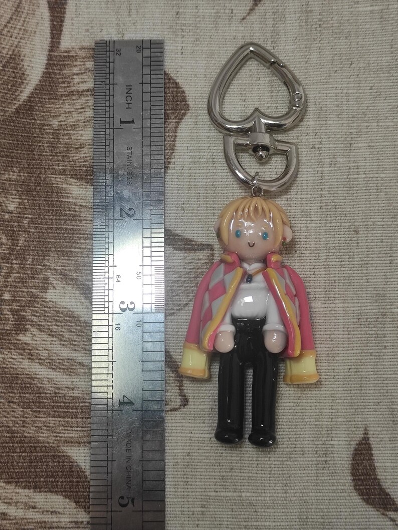 May include: A handmade keychain featuring a miniature figure of a character with blonde hair, blue eyes, and a pink and yellow jacket. The figure is attached to a silver-colored heart-shaped clasp. The figure is approximately 2 inches tall.
