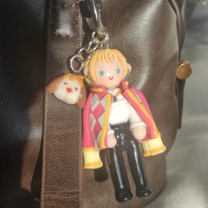 May include: A keychain featuring a miniature figurine of a character with blonde hair, wearing a pink and white patterned coat, black pants, and a white shirt. A small dog charm is also attached. The keychain is hanging from a silver chain.