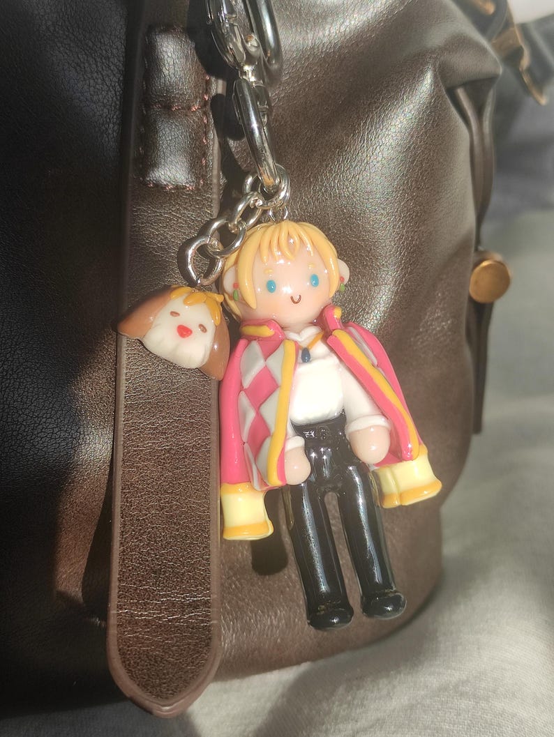 May include: A keychain featuring a miniature figurine of a character with blonde hair, wearing a pink and white patterned coat, black pants, and a white shirt. A small dog charm is also attached. The keychain is hanging from a silver chain.