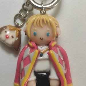 May include: A handmade keychain featuring a character from a popular animated film. The figure has blonde hair, blue eyes, and a pink and white checkered coat. A small dog charm hangs next to the main figure. The keychain is attached to a silver metal ring.