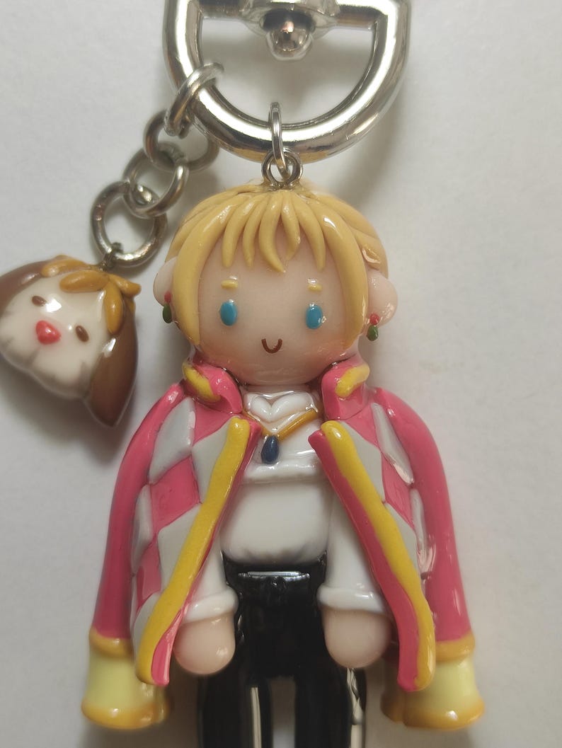 May include: A handmade keychain featuring a character from a popular animated film. The figure has blonde hair, blue eyes, and a pink and white checkered coat. A small dog charm hangs next to the main figure. The keychain is attached to a silver metal ring.
