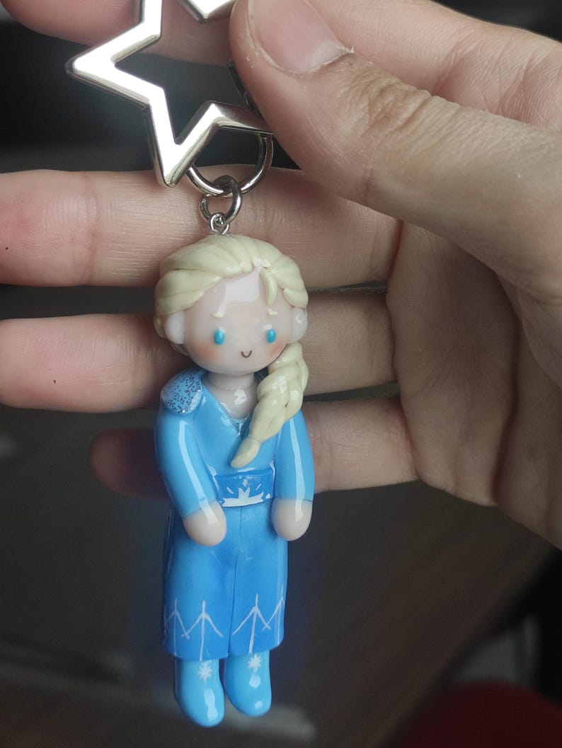 May include: A keychain featuring a miniature figurine of a character with blonde hair in a blue dress. The figurine has a silver star-shaped charm attached to the top. The character's dress has a white belt and snowflake details.