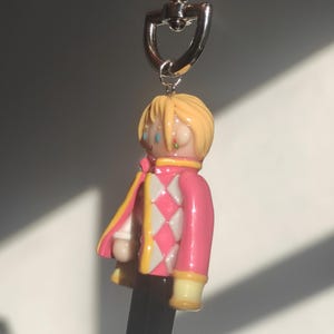 May include: A keychain featuring a miniature figure of a character with blonde hair, wearing a pink jacket with white and pink diamond pattern, black pants, and yellow accents. The keychain has a silver heart-shaped clasp.