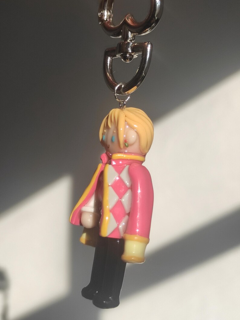 May include: A keychain featuring a miniature figure of a character with blonde hair, wearing a pink jacket with white and pink diamond pattern, black pants, and yellow accents. The keychain has a silver heart-shaped clasp.