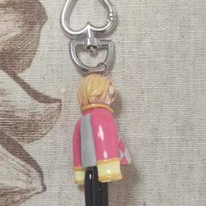May include: A keychain featuring a small figurine of a person with blonde hair, wearing a pink and gray jacket with yellow accents, black pants, and black boots. The keychain has a silver heart-shaped clasp.