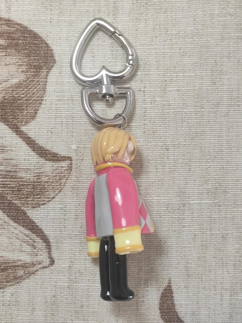 May include: A keychain featuring a small figurine of a person with blonde hair, wearing a pink and gray jacket with yellow accents, black pants, and black boots. The keychain has a silver heart-shaped clasp.