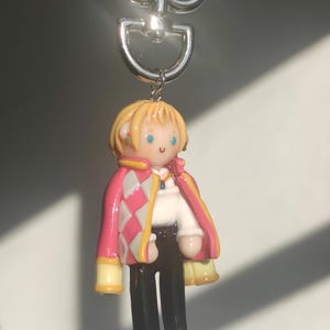 May include: A keychain featuring a miniature figure of a character with blonde hair, wearing a pink and white diamond-patterned jacket, a white shirt, and black pants. The figure has a silver-colored metal keyring.