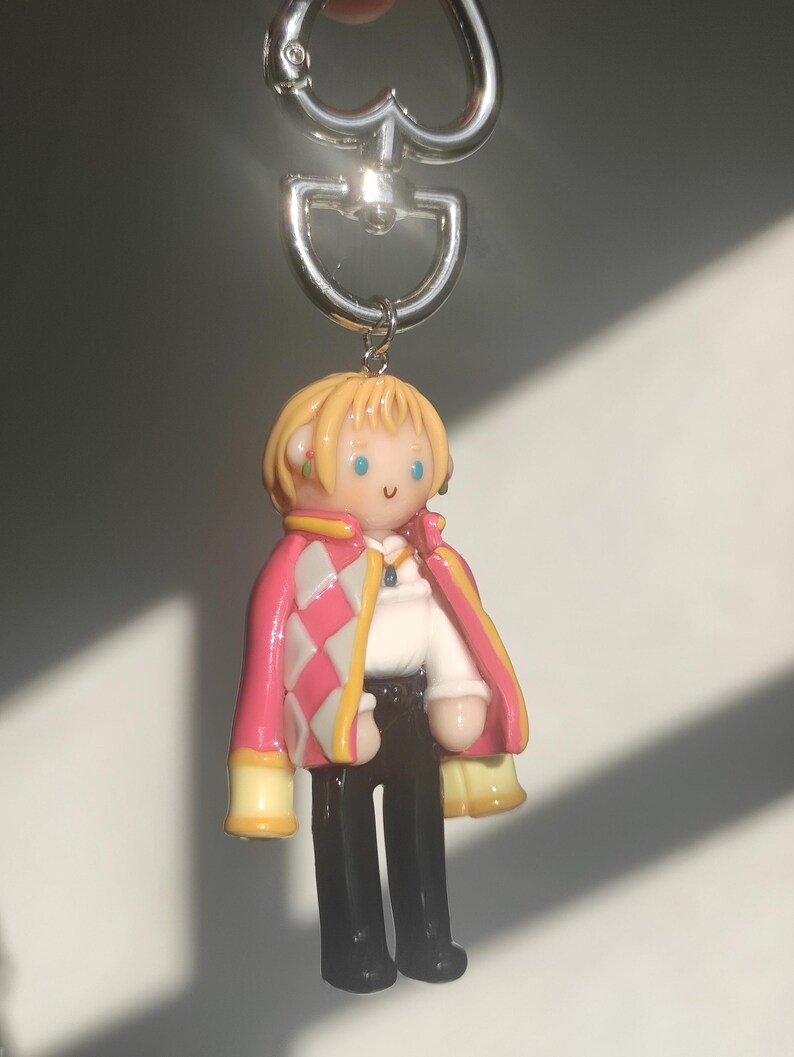 May include: A keychain featuring a miniature figure of a character with blonde hair, wearing a pink and white diamond-patterned jacket, a white shirt, and black pants. The figure has a silver-colored metal keyring.