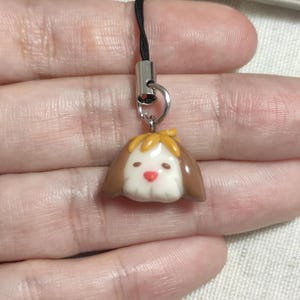 May include: A small, dog-shaped charm with a brown and white color scheme. The charm has a yellow bow and a red nose. It is attached to a black cord with a silver-colored metal clasp.