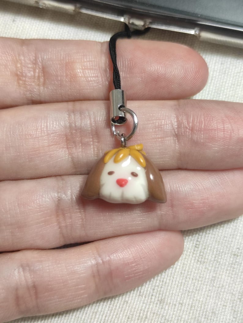 May include: A small, dog-shaped charm with a brown and white color scheme. The charm has a yellow bow and a red nose. It is attached to a black cord with a silver-colored metal clasp.