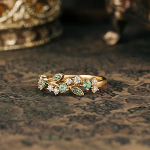 Puede incluir: Un anillo de oro con un diseño delicado que presenta una rama con piedras preciosas verdes y blancas. El anillo está colocado sobre una superficie texturizada de color marrón.