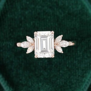 Op de afbeelding: Rosegouden verlovingsring met een grote, rechthoekige, heldere edelsteen als middelpunt. De ring wordt aan weerszijden geflankeerd door kleinere, marquise-geslepen stenen, waardoor een bloemachtig ontwerp ontstaat. De ring staat tegen een donkergroene achtergrond.