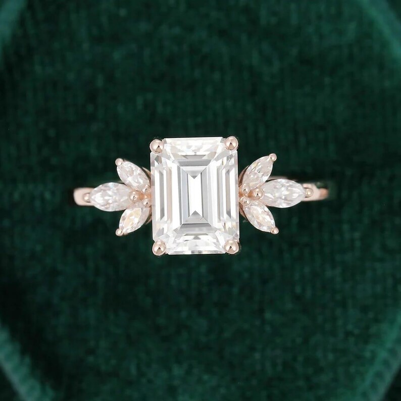 Op de afbeelding: Rosegouden verlovingsring met een grote, rechthoekige, heldere edelsteen als middelpunt. De ring wordt aan weerszijden geflankeerd door kleinere, marquise-geslepen stenen, waardoor een bloemachtig ontwerp ontstaat. De ring staat tegen een donkergroene achtergrond.