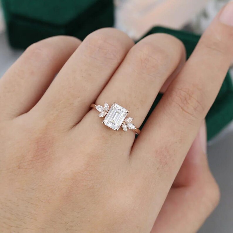 Op de afbeelding: Een ros&eacute;gouden ring met een heldere, smaragdgeslepen edelsteen als middelpunt. De ring is versierd met kleinere, marquise-geslepen stenen aan weerszijden, waardoor een bladachtig ontwerp ontstaat. De ring wordt op een hand getoond.