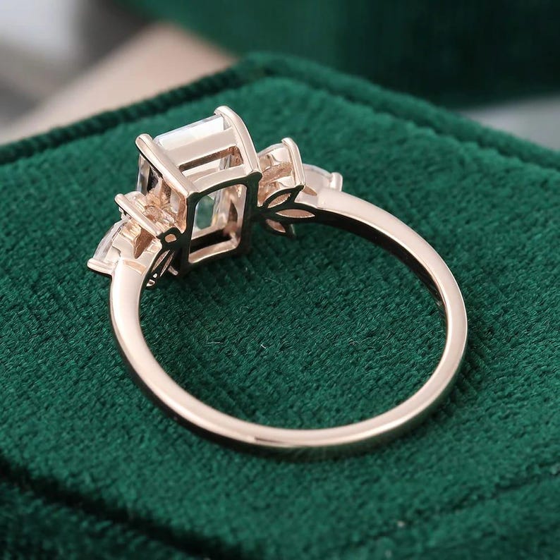 Op de afbeelding: Rosegouden verlovingsring met een grote, rechthoekig geslepen middensteen en kleinere, peervormige zijstenen. De ring wordt getoond op een donkergroene fluwelen achtergrond, die het ontwerp en de glans van de ring benadrukt.