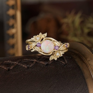 Könnte beinhalten: Ein goldener Ring mit einem weißen Opal-Edelstein, umgeben von zarten Blättern und violetten Amethyst-Akzenten.
