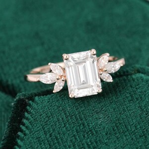 Op de afbeelding: Een ros&eacute;gouden ring met een grote, rechthoekige, heldere edelsteen als middelpunt. De ring is versierd met kleinere, marquise-geslepen stenen aan weerszijden van de hoofdsteen, waardoor een bloemmotief ontstaat. De ring staat tegen een donkergroene fluwelen achtergrond.