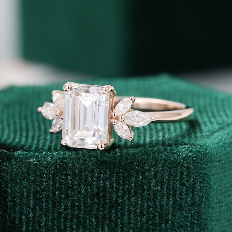 Op de afbeelding: Een ros&eacute;gouden verlovingsring met een smaragdgeslepen middensteen. De ring heeft aan weerszijden van de middensteen een cluster van marquise-geslepen stenen. De ring ligt op een donkergroen fluwelen oppervlak.