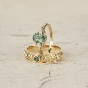 Op de afbeelding: Een paar gouden trouwringen met een groene edelsteen en een bladontwerp. De verlovingsring heeft een grote ovale groene edelsteen omringd door kleinere diamanten.