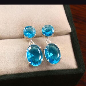Könnte beinhalten: Ein Paar silberne Ohrringe mit leuchtend blauen, oval geschliffenen Edelsteinen. Jeder Ohrring hat einen kleineren runden Stein oben und einen größeren ovalen Stein, der darunter hängt. Die Ohrringe werden in einer Schmuckschatulle präsentiert.