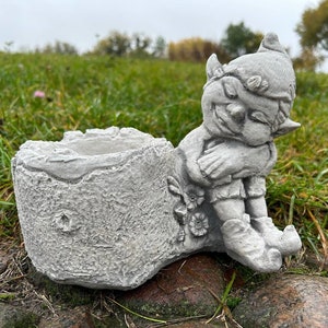 Op de afbeelding: Een grijze betonnen dwergpot met een slapende dwergfiguur die zijn armen vasthoudt. De pot is gevormd als een boomstronk met een klein gat om te planten.