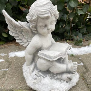 May include: A gray stone angel figurine with wings, sitting on a snowy base and holding a book. The angel has a sweet expression and is wearing a simple dress.
