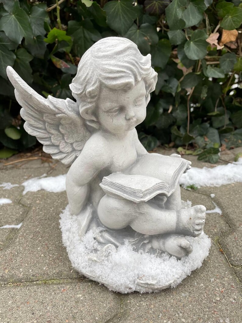 May include: A gray stone angel figurine with wings, sitting on a snowy base and holding a book. The angel has a sweet expression and is wearing a simple dress.