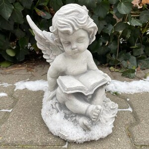 May include: A white stone angel figurine with wings, sitting on a base covered in faux snow, holding an open book.