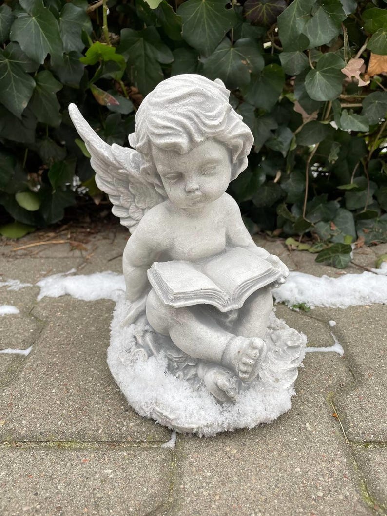 May include: A white stone angel figurine with wings, sitting on a base covered in faux snow, holding an open book.