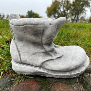 Peut inclure: Un pot de fleurs en béton gris en forme de botte. La botte est patinée et a un aspect usé. Elle est posée sur un lit de gravier.