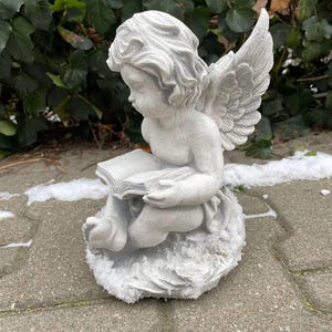 May include: A white stone angel figurine with wings, sitting on a stone base, holding a book in its hands.