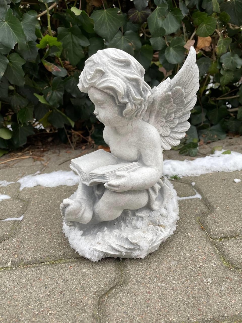 May include: A white stone angel figurine with wings, sitting on a stone base, holding a book in its hands.