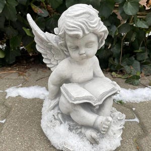 May include: A gray concrete cherub statue sitting on a base covered in faux snow. The cherub is holding a book and has wings.