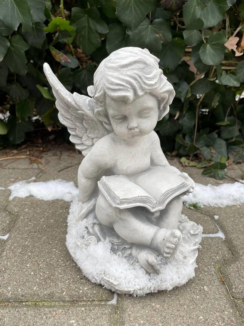 May include: A gray concrete cherub statue sitting on a base covered in faux snow. The cherub is holding a book and has wings.