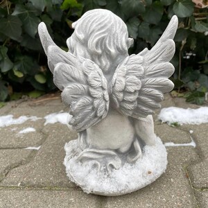 May include: A gray concrete angel statue with wings, sitting on a base covered in white snow. The angel has its head tilted down and its wings are spread out behind it.