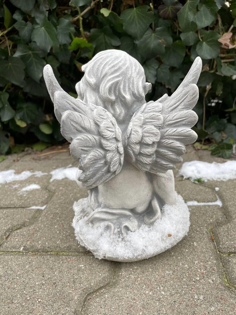 May include: A gray concrete angel statue with wings, sitting on a base covered in white snow. The angel has its head tilted down and its wings are spread out behind it.